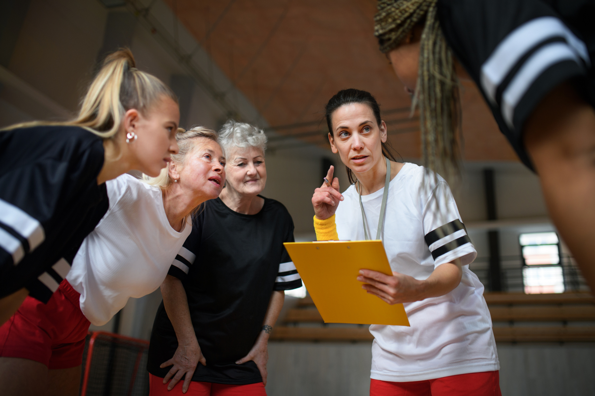 Female sport coach with clipboard discussing tactics with young and old women team training for match in gym.