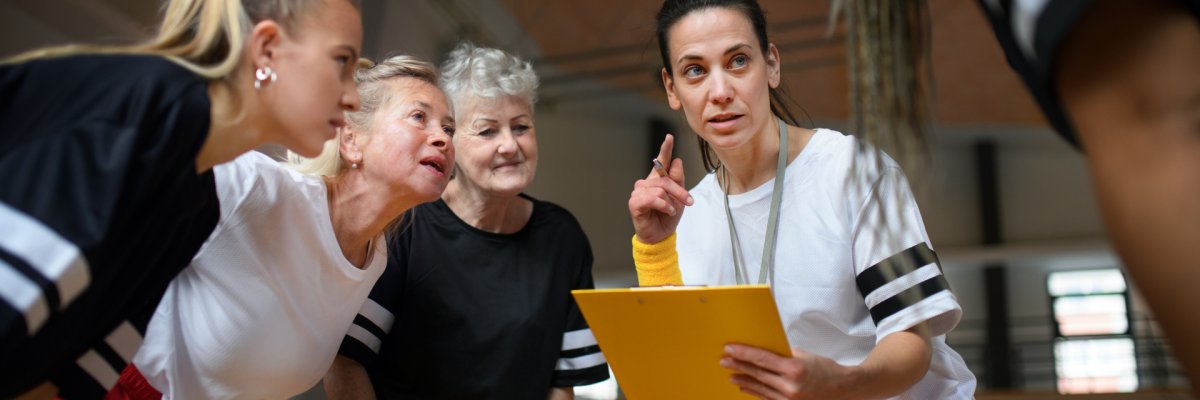 Female sport coach with clipboard discussing tactics with young and old women team training for match in gym.