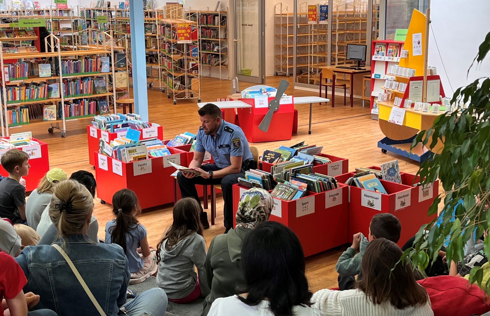Ein Polizist sitzt auf einem Stuhl in einer Bücherei und liest Kindern Geschichten vor.