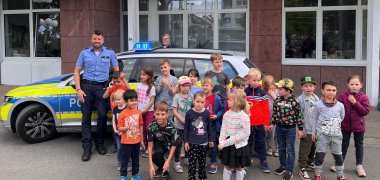 Ein Polizist steht lachend vor einem Polizeiauto und einer Gruppe von Kindern.