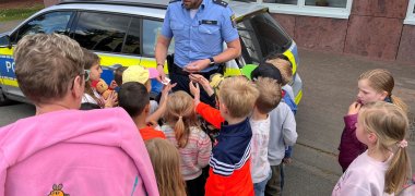 Ein Polizist verteilt kleine Aufkleber an eine Gruppe von Kindern. Im Hintergrund steht ein Polizeiauto.