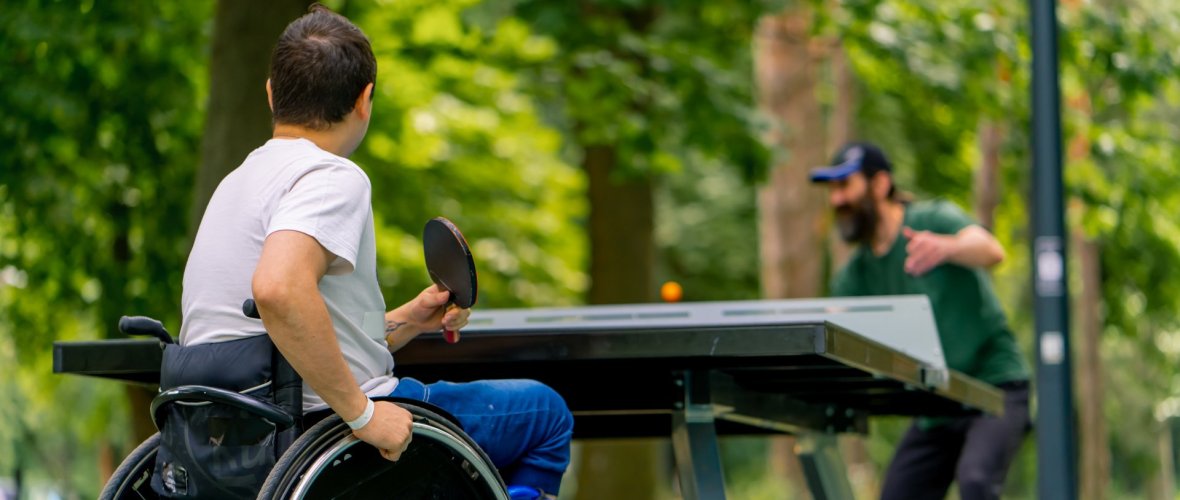 A disabled man in a wheelchair plays ping pong with an older man in a city park against backdrop of trees A disabled man in a wheelchair plays ping pong with an older man in a city park against backdrop of trees